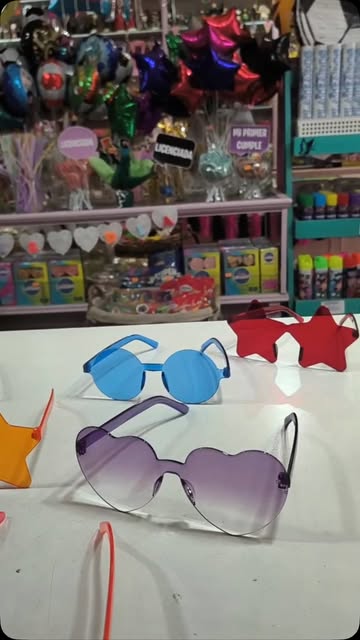 Anteojos de colores con forma de corazón y estrella sobre una mesa, con globos metalizados al fondo en el local de Cotillón Cotipel