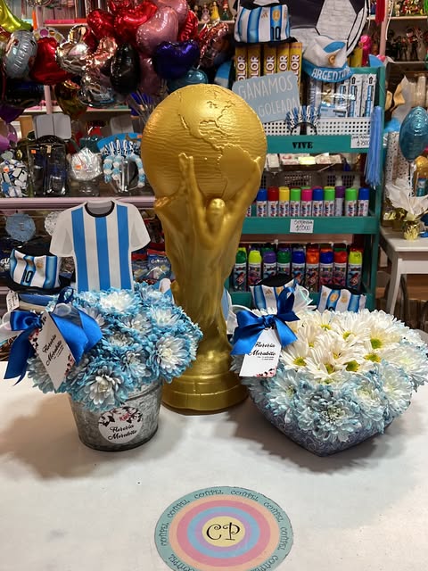 Copa dorada decorativa, camiseta celeste y blanca y arreglos florales en celeste sobre una mesa del local